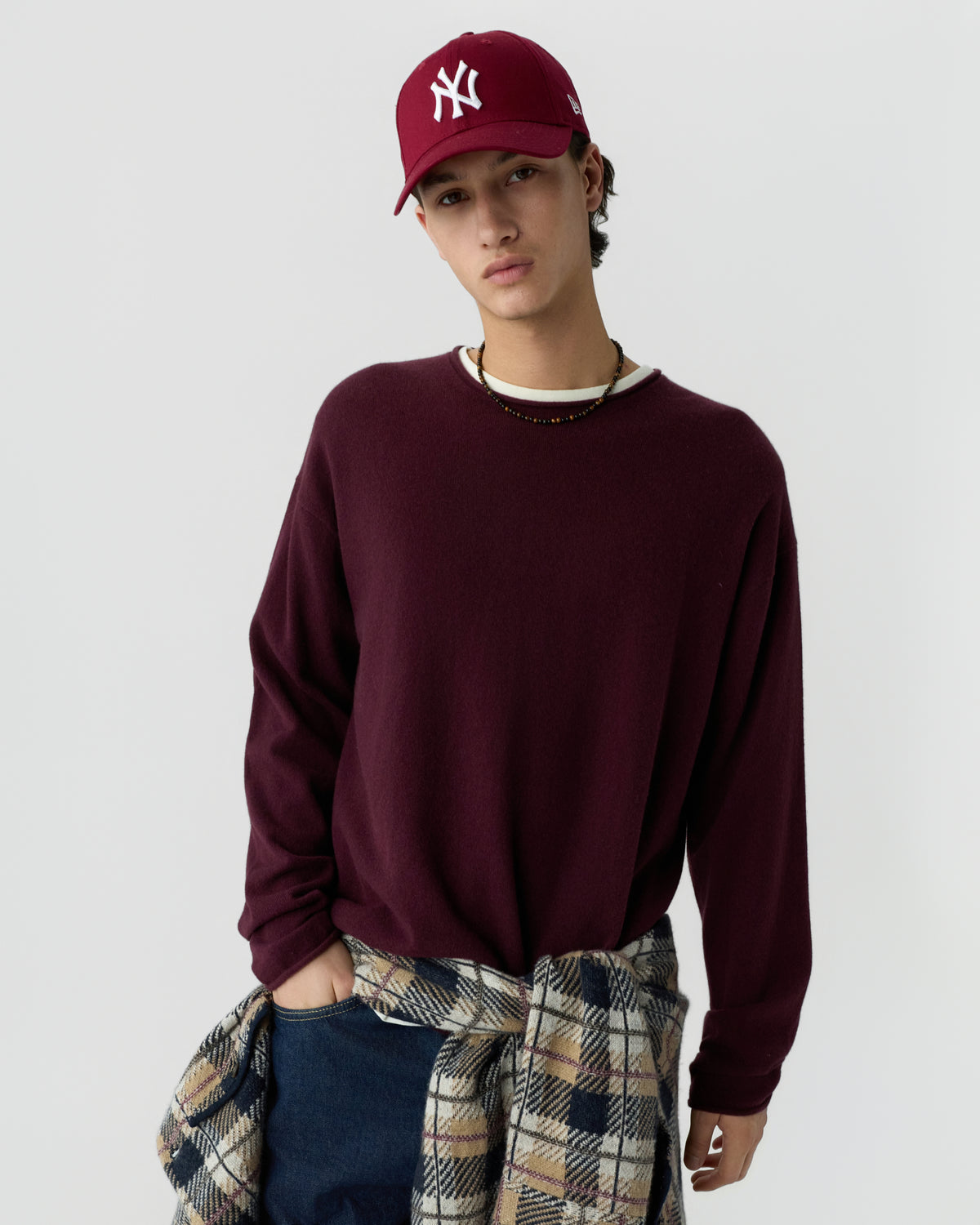 Young man wearing a burgundy cashmere sweater and matching baseball cap, with a plaid shirt tied around his waist against a plain light background.