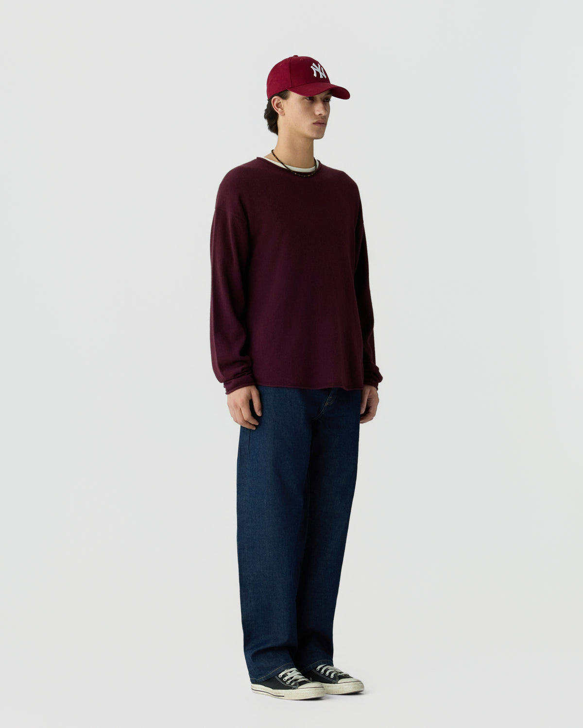 A young man standing against a plain light background wearing a maroon long-sleeve sweater, dark blue jeans, black sneakers, a maroon baseball cap with white lettering, and a necklace.