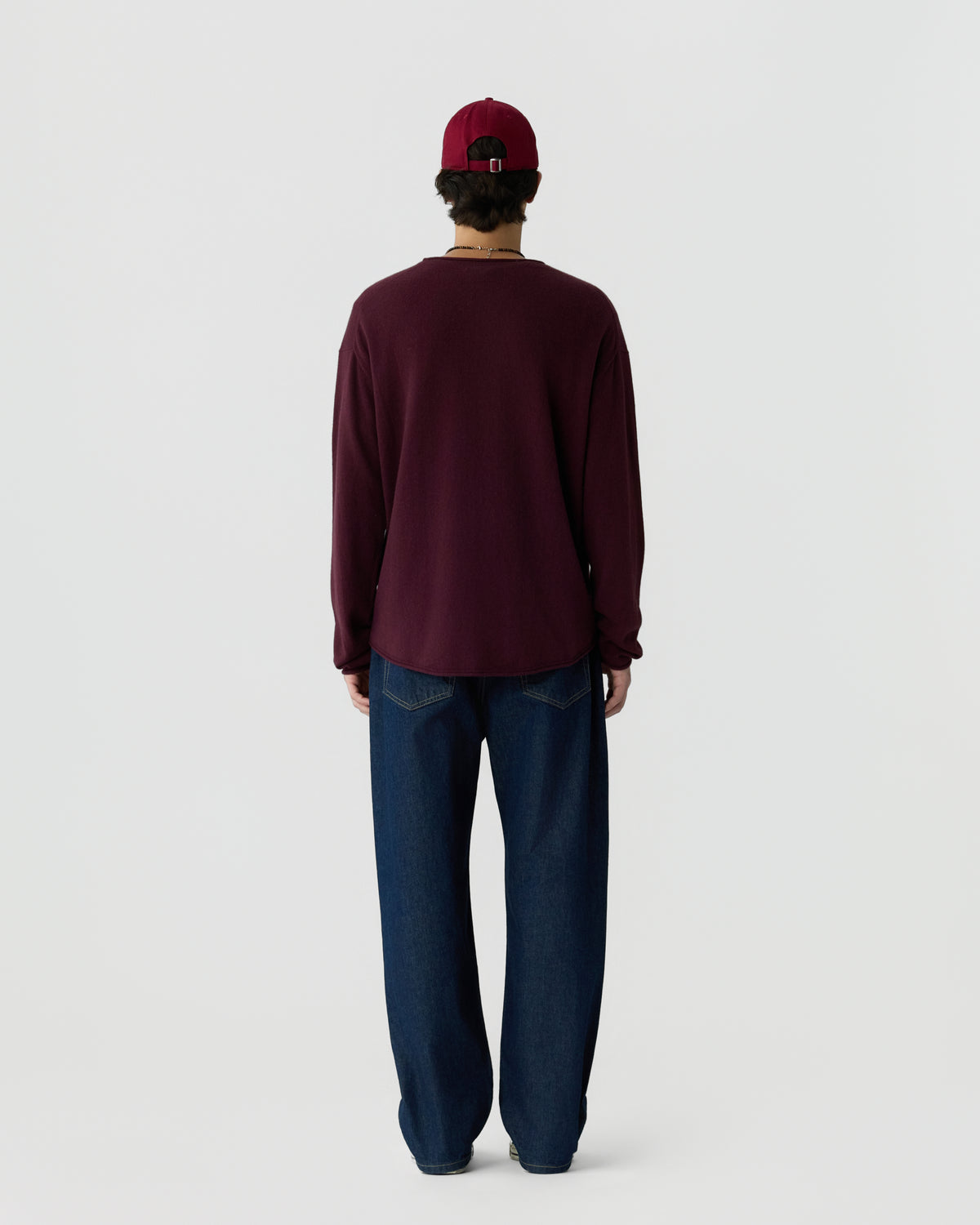 Man wearing a burgundy long-sleeve shirt, dark blue jeans, and a matching burgundy baseball cap, standing facing away against a plain light background.