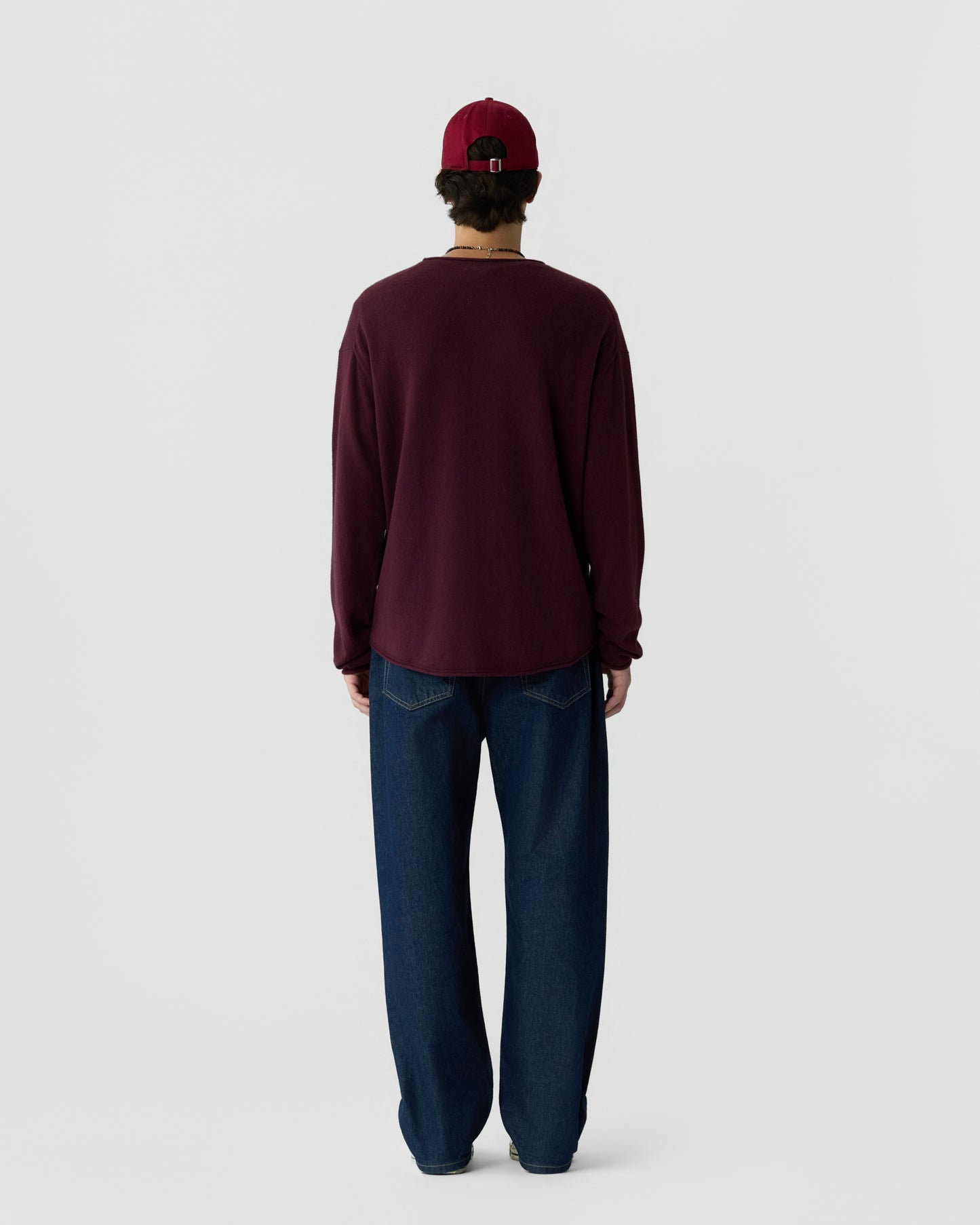 Man wearing a burgundy long-sleeve shirt, dark blue jeans, and a matching burgundy baseball cap, standing facing away against a plain light background.