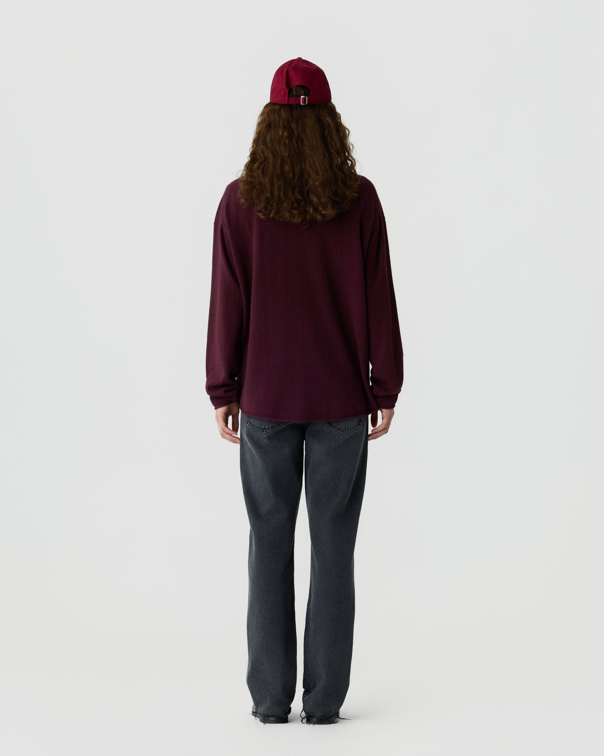 Person wearing a burgundy long-sleeve shirt, dark gray jeans, and a matching burgundy baseball cap, standing and facing away against a plain white background.