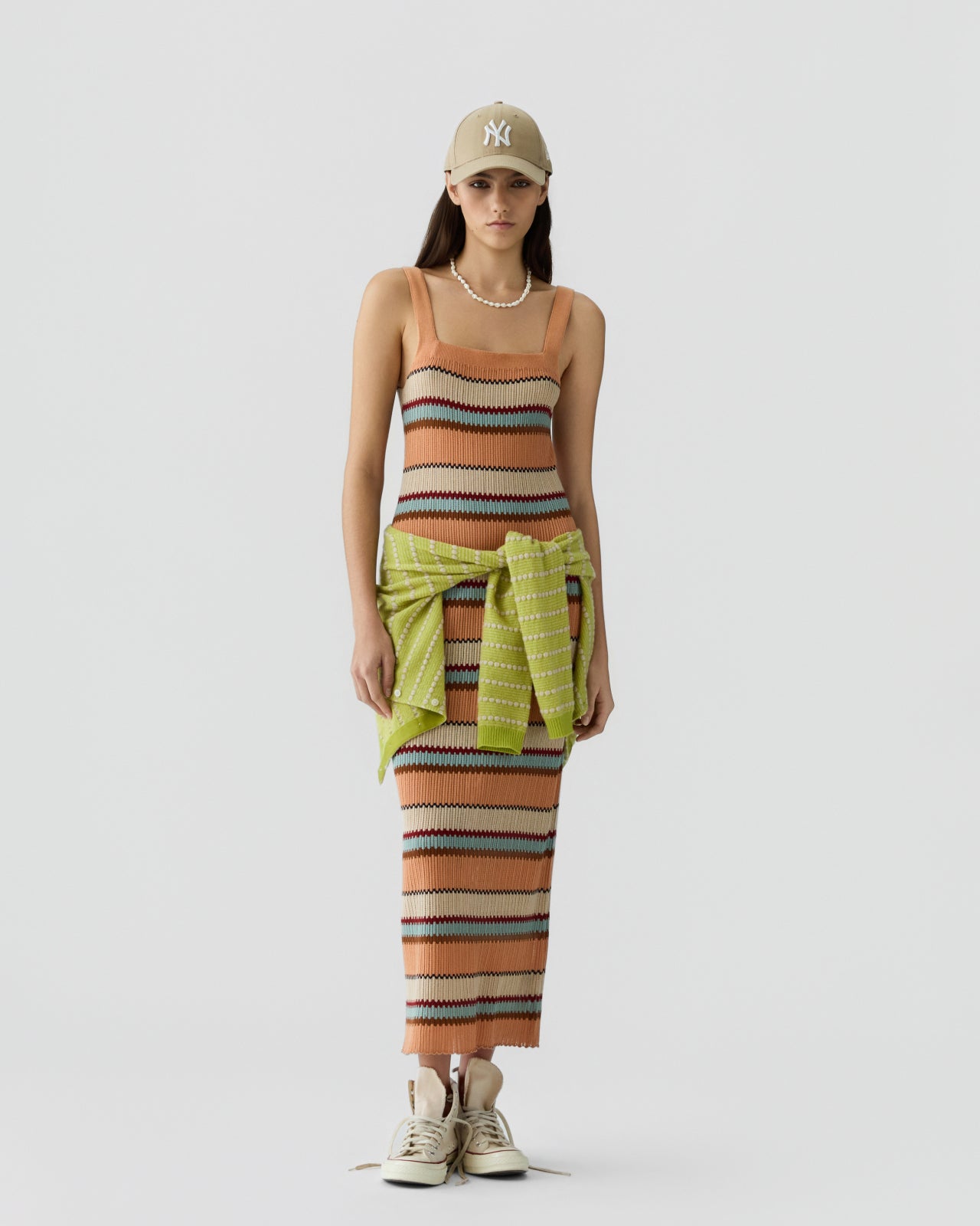 Woman wearing a sleeveless, ribbed knit dress with horizontal stripes in warm tones of brown, beige, teal, and rust, with a light green sweater tied around her waist and beige sneakers, paired with a beige baseball cap against a plain background.