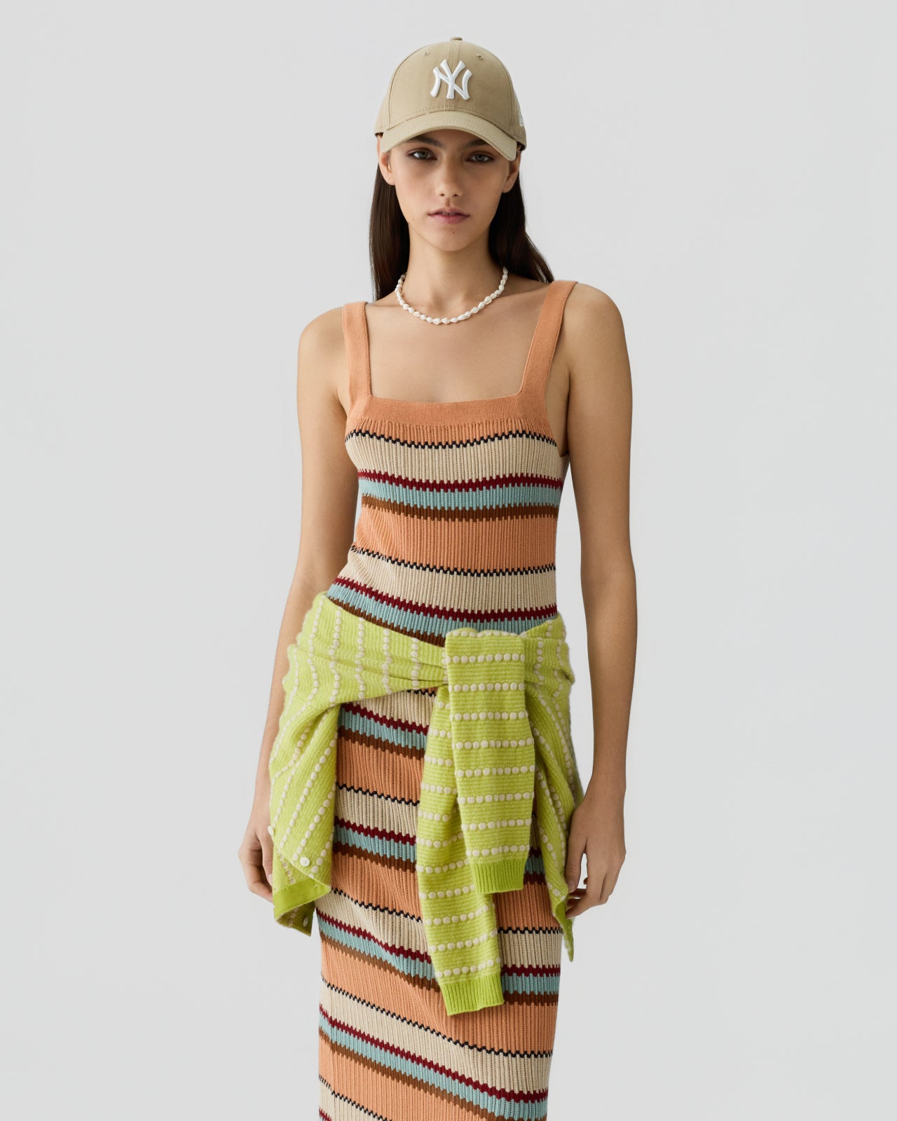 Young woman wearing a sleeveless knit dress with horizontal stripes in shades of orange, beige, brown, and teal, accessorized with a pale green sweater tied around her waist, a pearl necklace, and a beige baseball cap against a plain light background.
