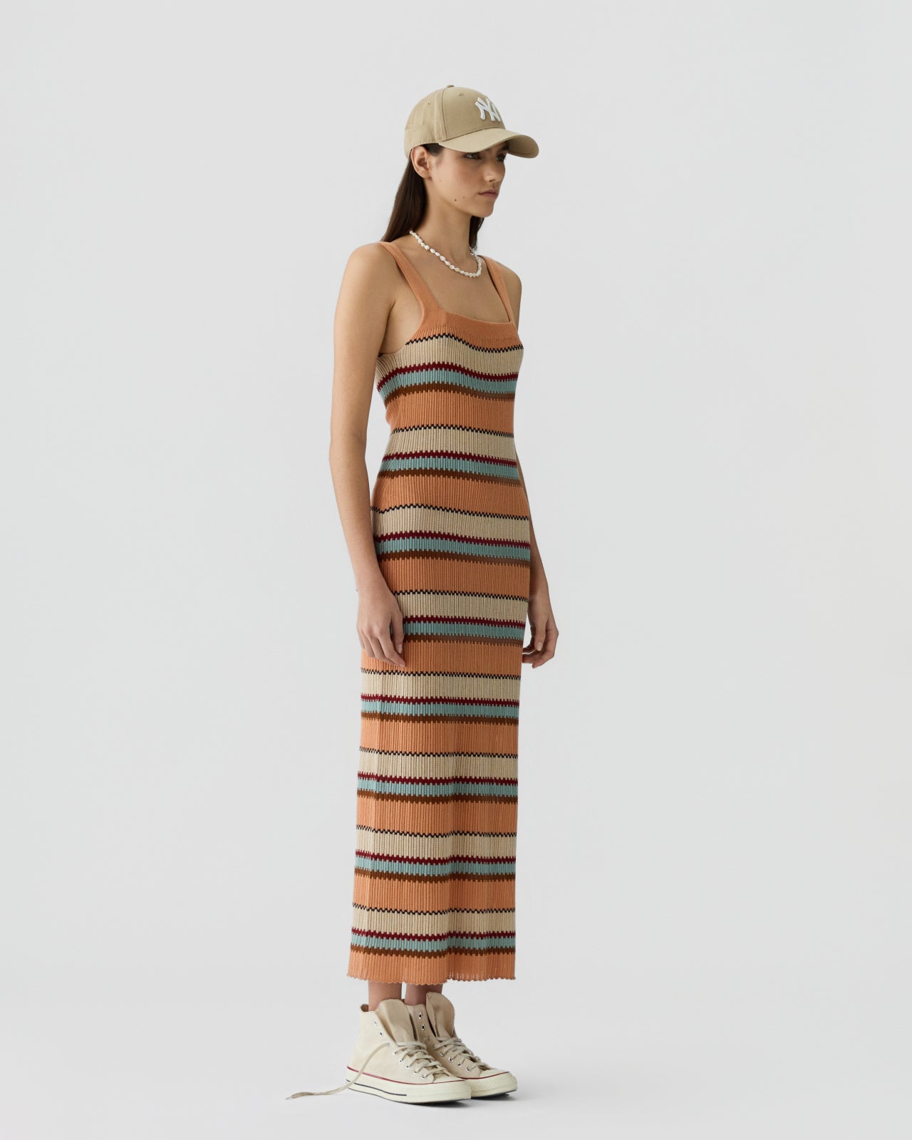 Slim woman with long dark hair wearing a sleeveless knit dress with horizontal stripes in shades of orange, tan, brown, and blue, paired with high-top sneakers and a beige baseball cap against a plain white background.