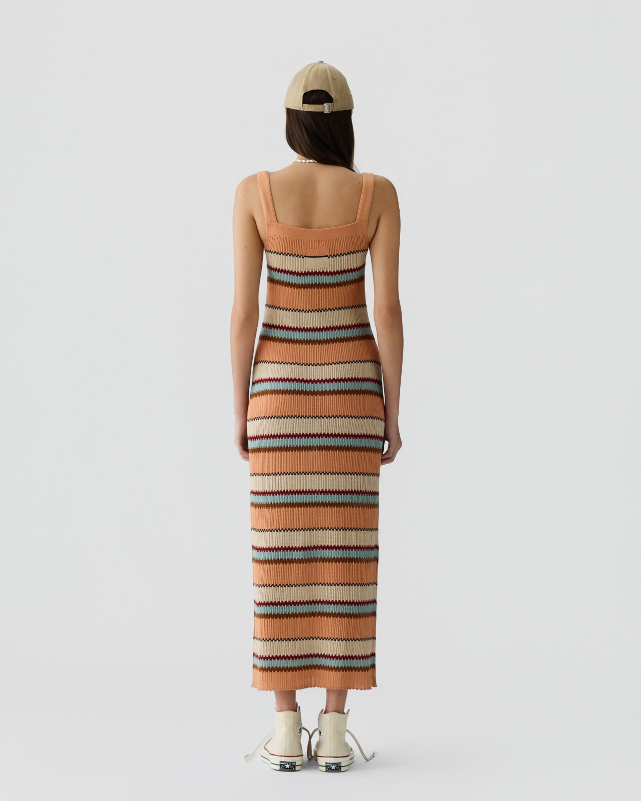 Woman with long dark hair wearing a sleeveless knit dress with narrow horizontal stripes in warm tones of peach, cream, teal, and brown, paired with a beige cap and light sneakers, standing against a plain background.