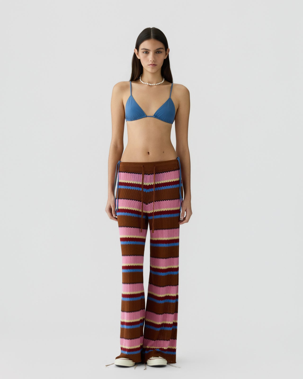 Young woman with straight dark hair wearing relaxed-fit knit pants featuring horizontal stripes in shades of brown, pink, blue, and cream paired with a blue bralette and shell necklace against a plain gray background.