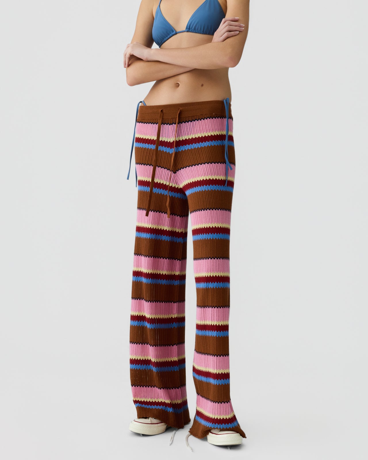 Woman standing with arms crossed wearing wide-leg knit pants with horizontal stripes in brown, pink, blue, and cream tones paired with a blue triangle bikini top against a plain background.