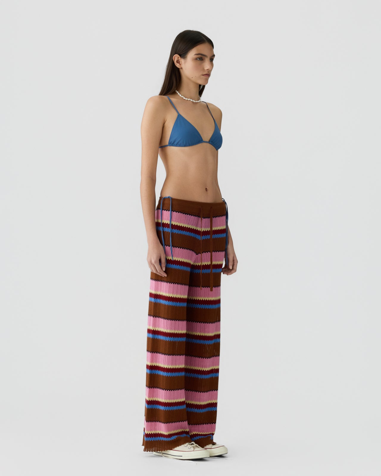 Slim woman with long dark hair wearing wide-leg knit pants with horizontal stripes in brown, pink, blue, and cream, paired with a blue triangle bikini top and shell necklace against a plain background.