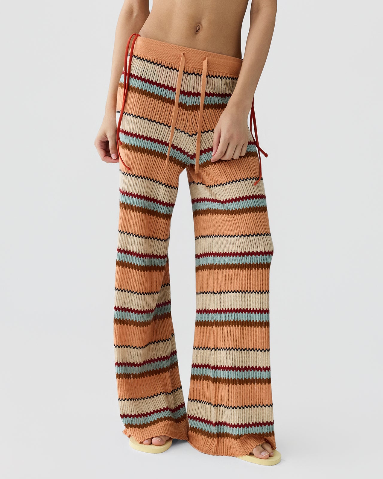 Person wearing high-waisted knit pants with horizontal stripes in warm tones of orange, cream, brown, and light blue, featuring a drawstring waist and wide legs against a plain background.