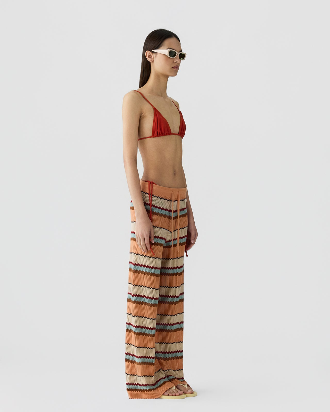 Slim woman with straight dark hair wearing loose knit wide-leg pants with horizontal zigzag stripes in warm shades of orange, cream, and black, paired with a red bikini top and white sunglasses against a plain background.