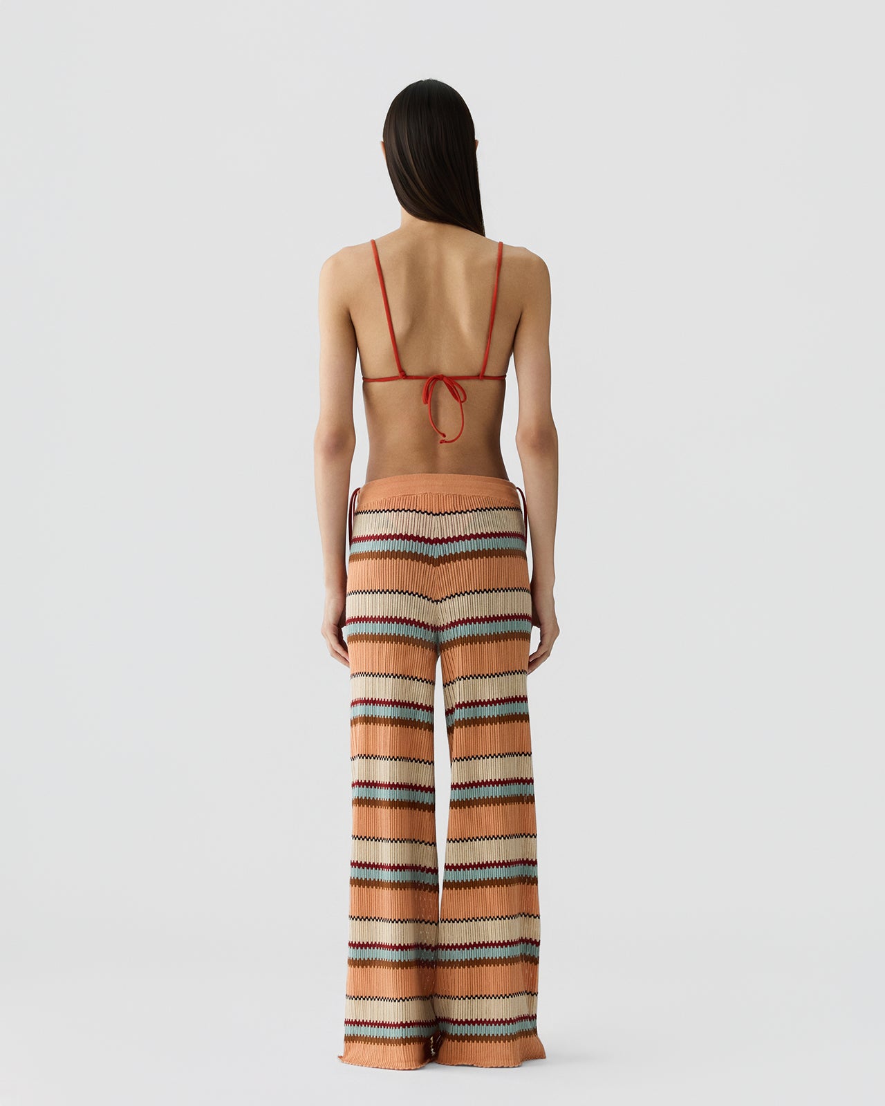 Woman with long dark hair seen from behind wearing wide-leg knit pants with horizontal stripes in shades of peach, brown, cream, and teal, paired with a thin-strapped orange bikini top against a plain background.