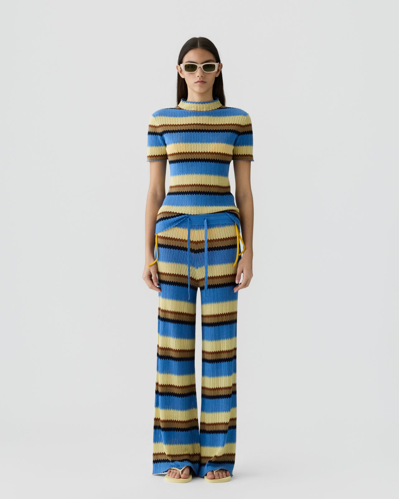 Woman with straight dark hair wearing a ribbed knit short-sleeve top and matching wide-leg pants featuring horizontal stripes in shades of blue, tan, cream, and brown, accessorized with light-colored sunglasses against a plain light background.