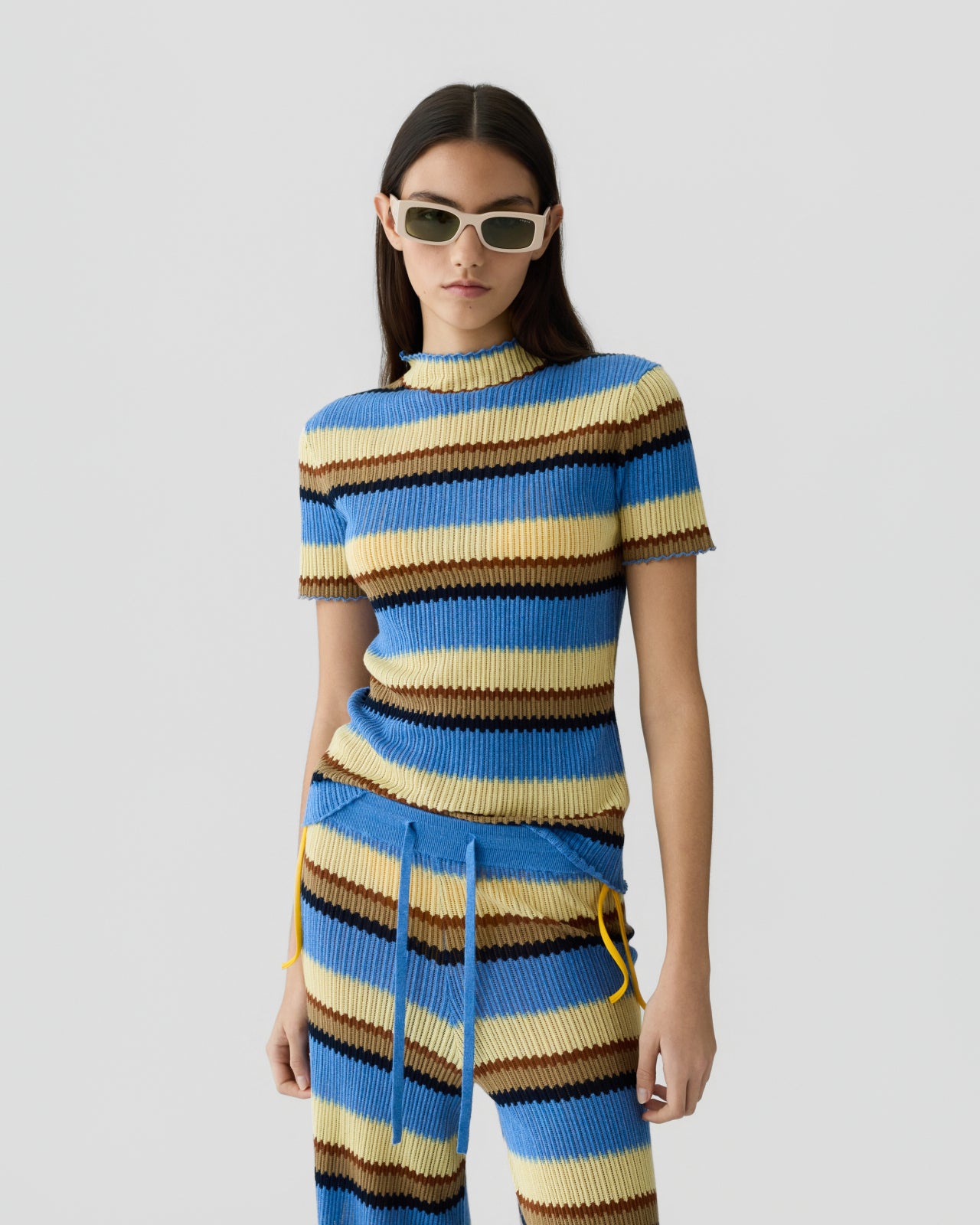 Woman with straight dark hair wearing a ribbed knit short-sleeve top and matching drawstring pants with horizontal stripes in shades of blue, yellow, cream, and brown, paired with rectangular sunglasses against a plain light background.