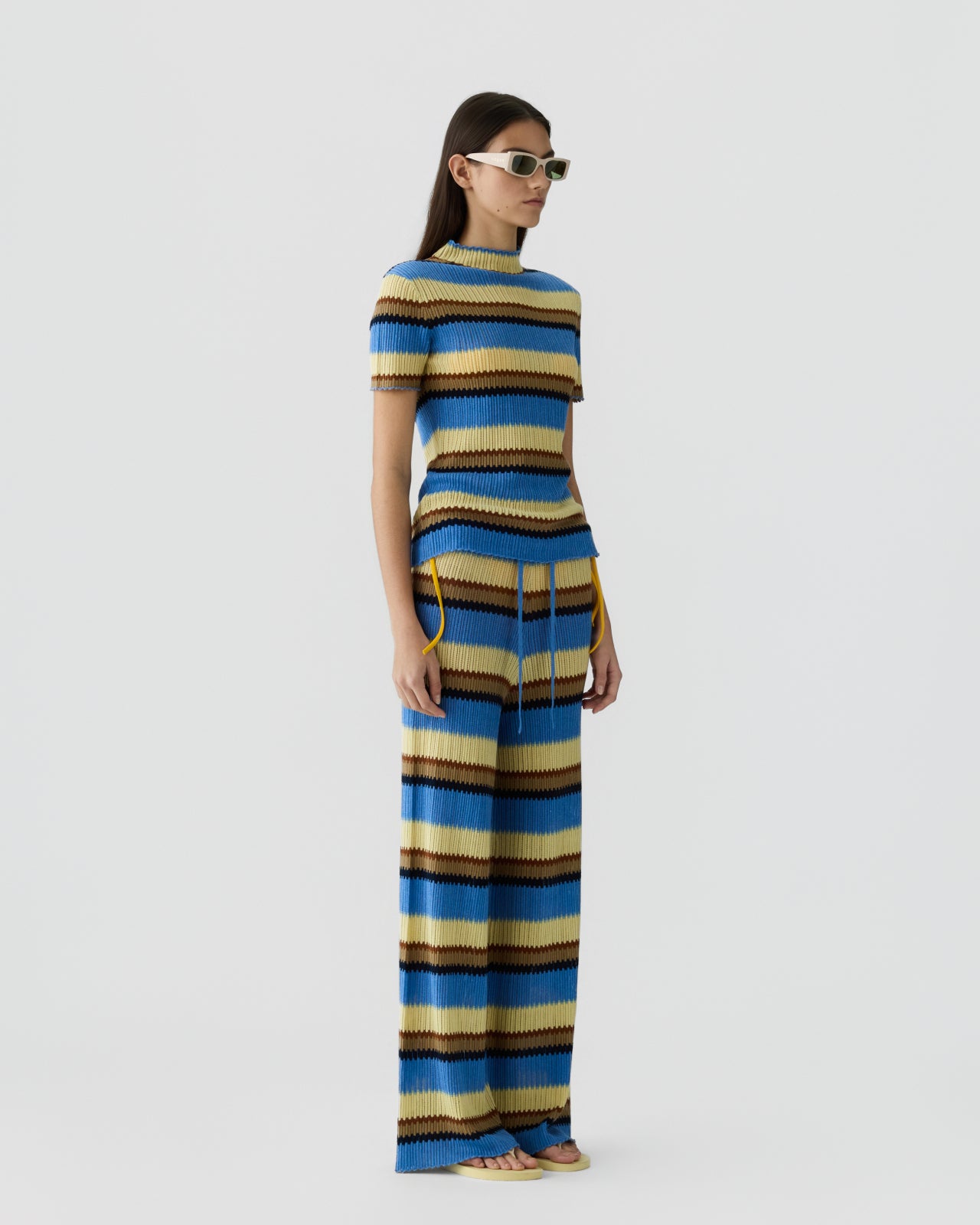 Woman with straight dark hair wearing a ribbed knit short-sleeve top and matching wide-leg pants with horizontal stripes in shades of blue, tan, cream, and brown, paired with narrow sunglasses against a plain light background.