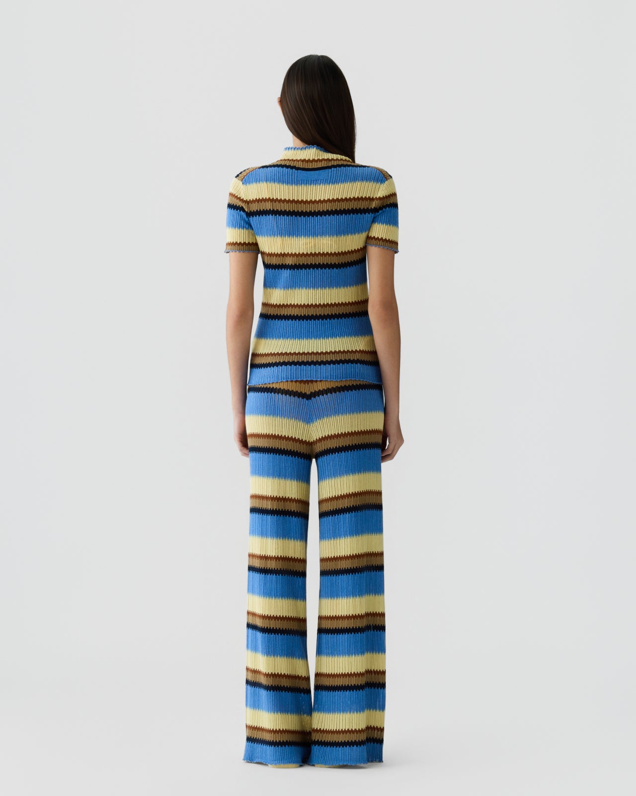 Woman with straight dark hair seen from behind wearing a ribbed knit short-sleeve top and wide-leg pants with horizontal stripes in shades of blue, tan, black, and cream against a plain background.