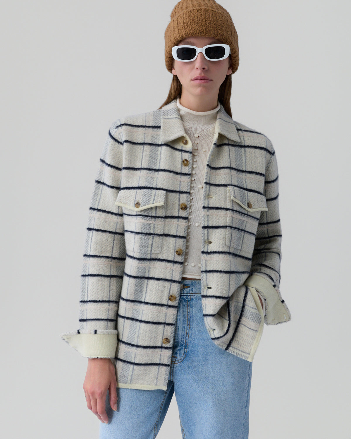 Person wearing an off-white plaid wool shirt jacket with navy stripes over a cream textured sweater, paired with light blue jeans, white rectangular sunglasses, and a brown knit beanie against a plain light background.