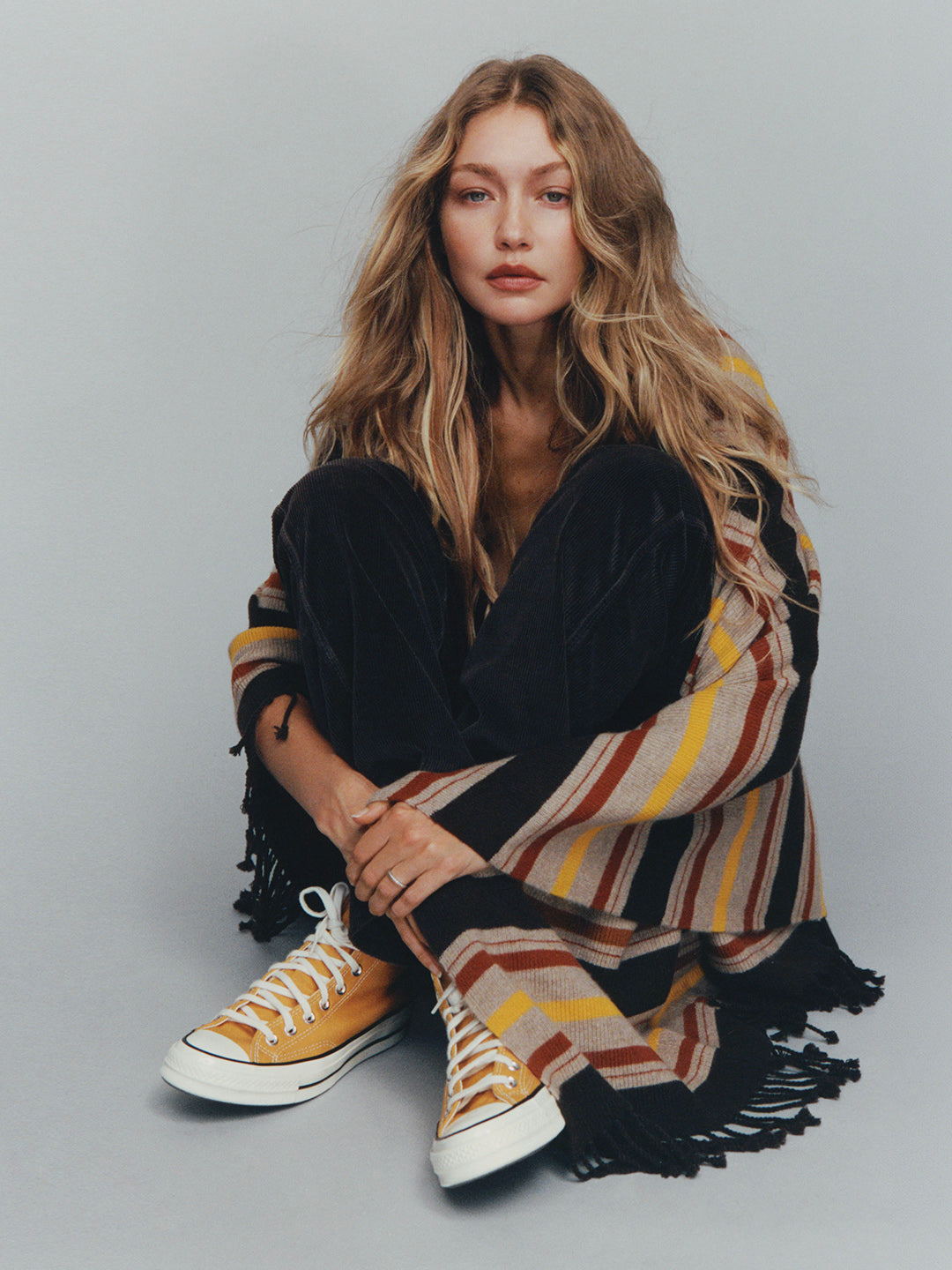 Woman seated on the floor wearing a striped fringed cardigan black pants and white sneakers against a gray background