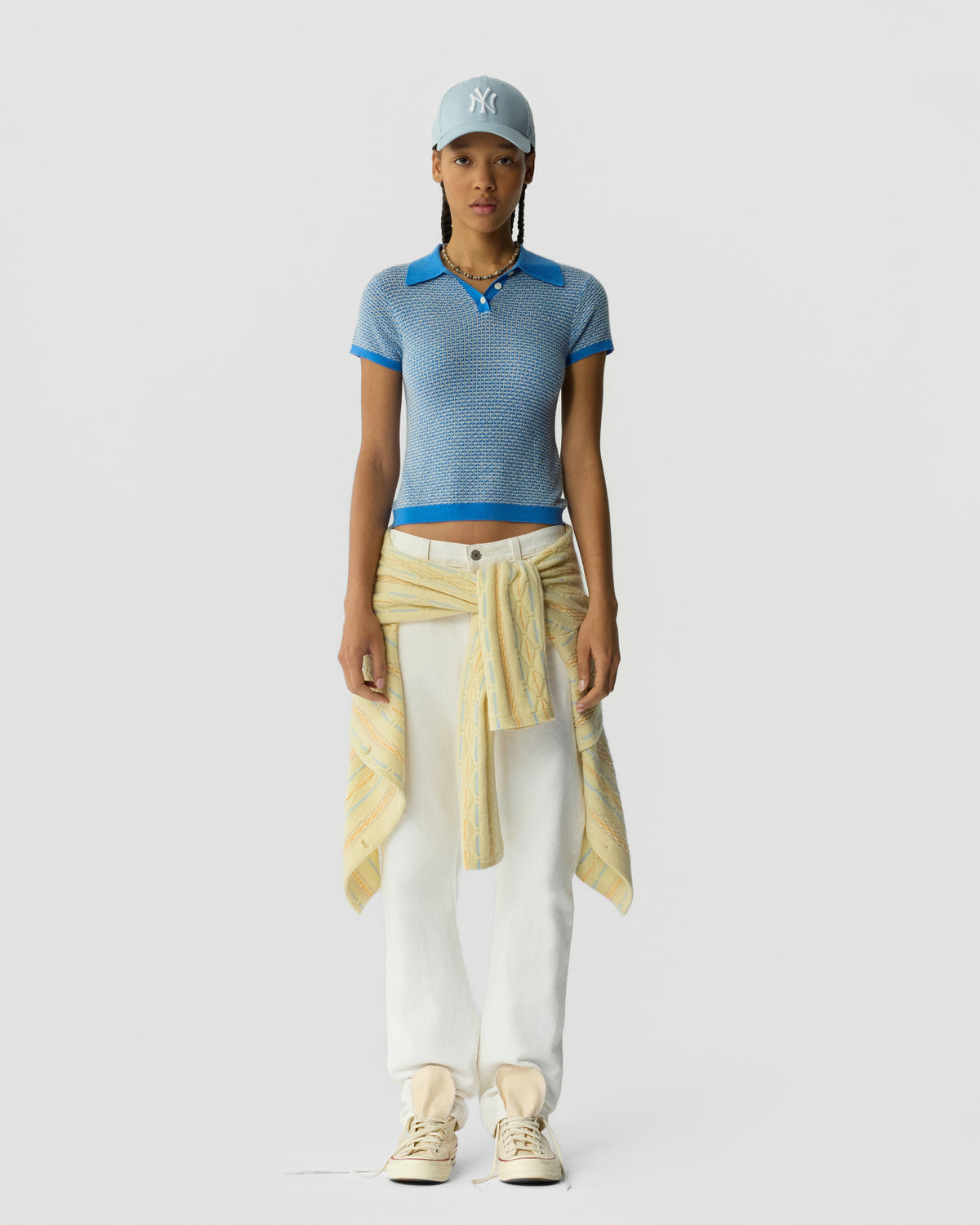 Woman standing facing forward wearing a fitted short-sleeve knit polo in a blue textured pattern with solid bright blue collar and sleeve trim, paired with straight white pants and a light blue baseball cap, with a yellow patterned sweater tied around the waist and cream sneakers.