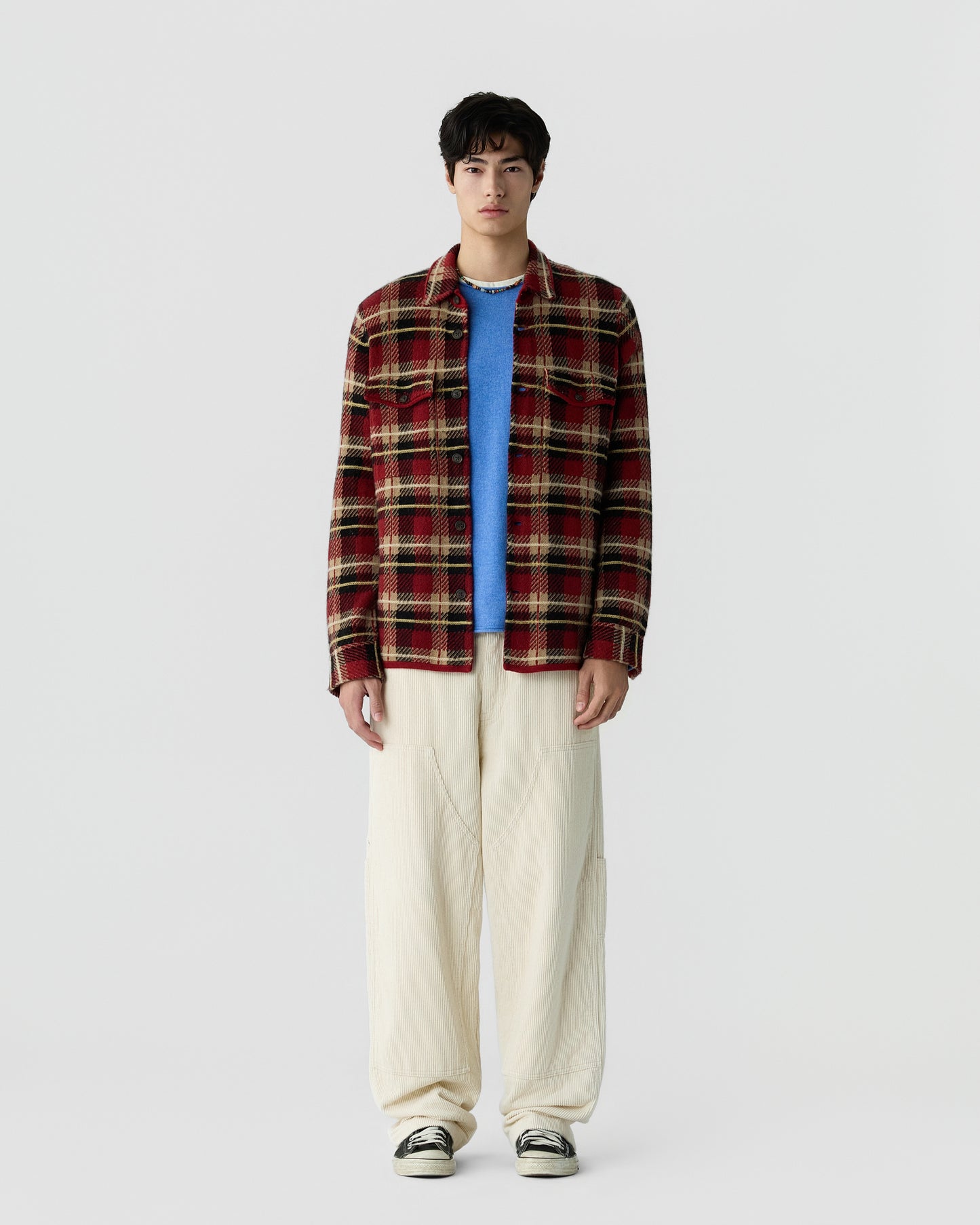 A young man stands wearing a red and brown plaid jacket over a blue shirt, loose cream-colored pants, and black and white sneakers against a plain white background.