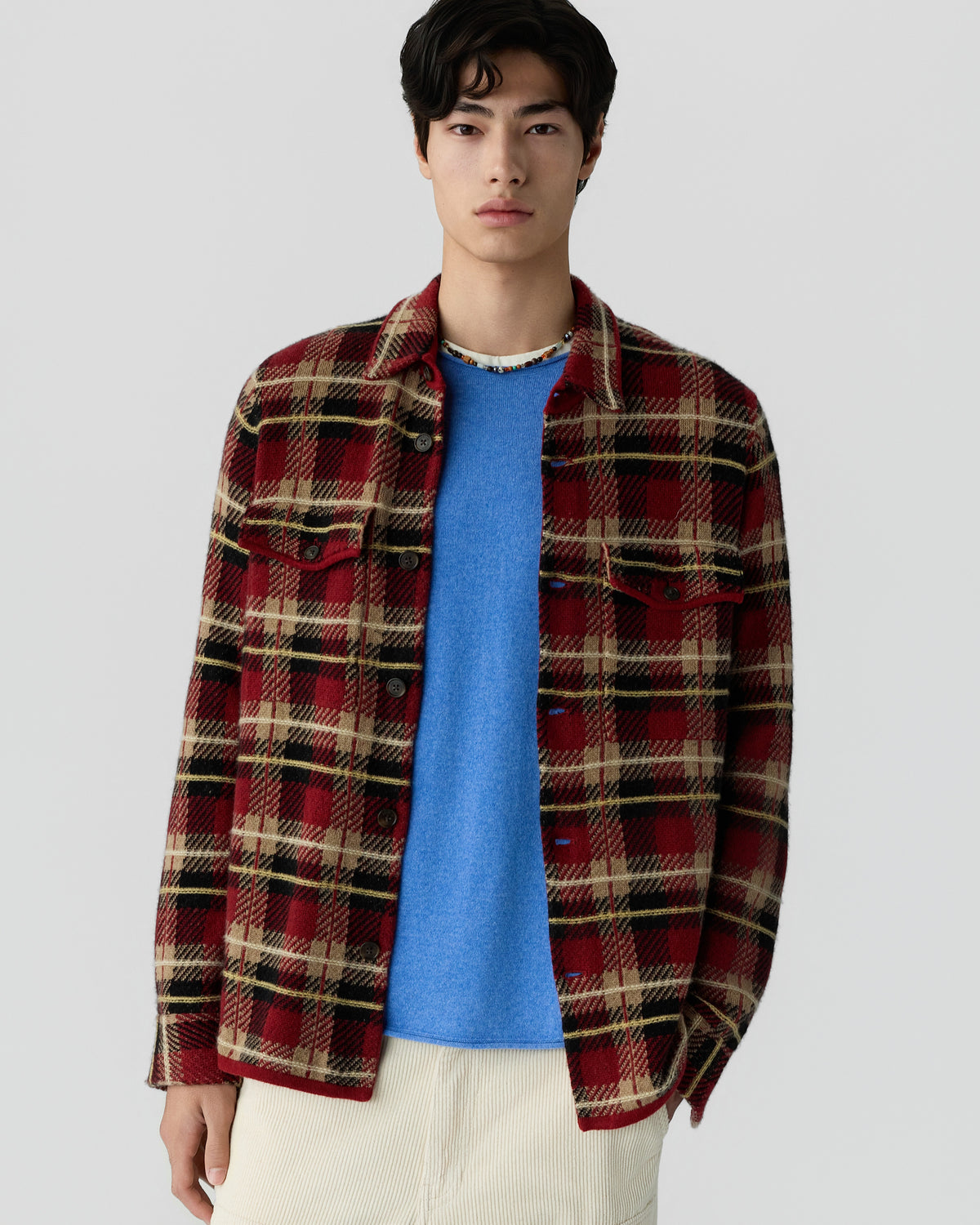 Young man wearing a red and beige plaid button-up shirt over a bright blue sweater with cream-colored pants against a plain light gray background.