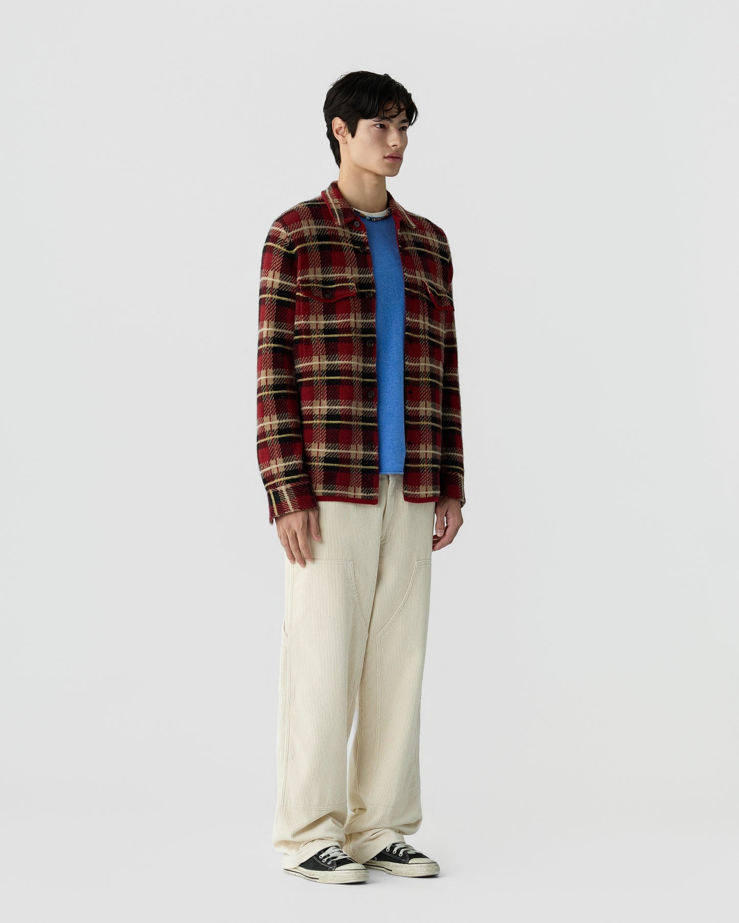 Young man wearing a red and beige plaid jacket over a bright blue sweater, paired with loose cream-colored pants and black sneakers, standing against a plain gray background.