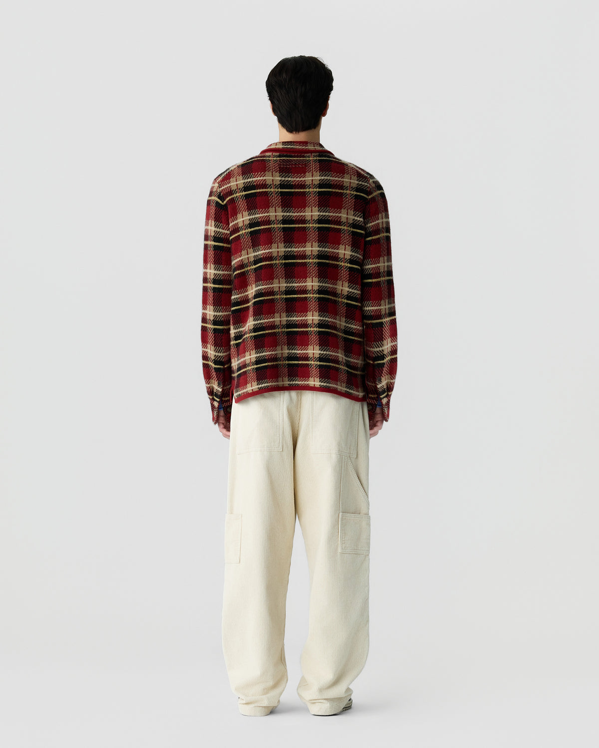 Man standing facing away wearing a red and black plaid long-sleeve shirt and loose-fitting off-white pants against a plain white background.