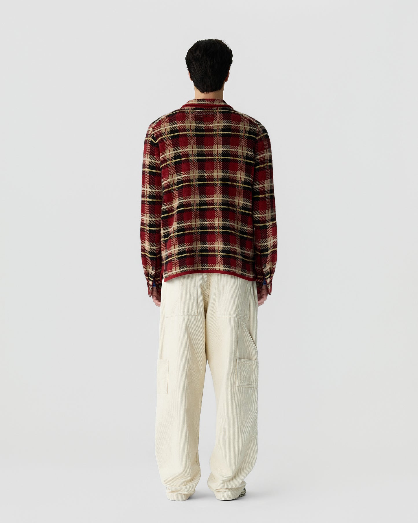 Man standing facing away wearing a red and black plaid long-sleeve shirt and loose-fitting off-white pants against a plain white background.