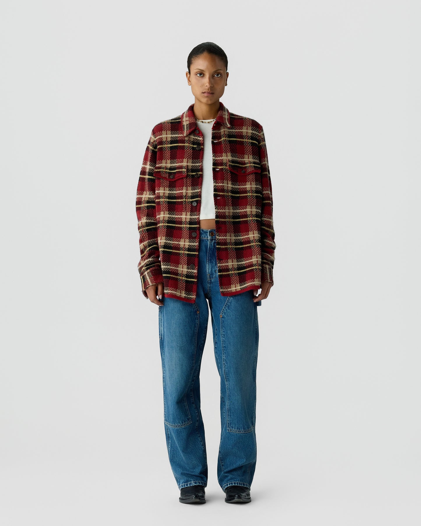 A woman stands facing forward wearing a red and black plaid button-up shirt over a white cropped top, paired with loose blue jeans and black shoes on a plain white background.