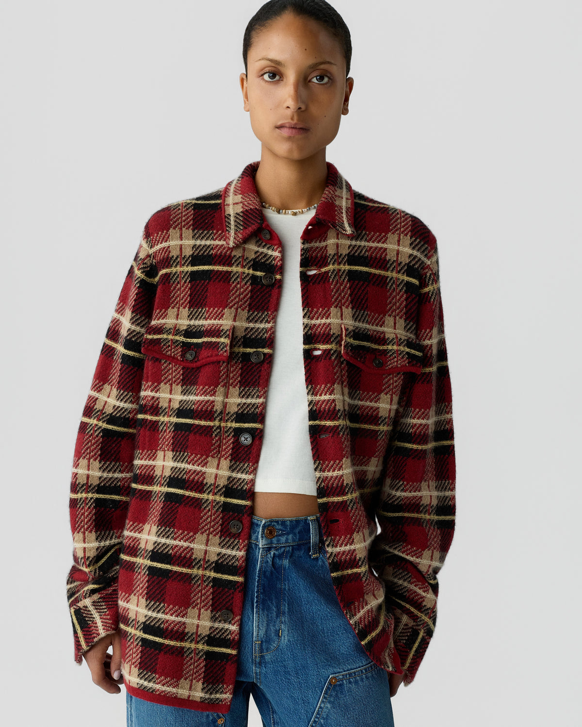 Woman wearing a red, black, and beige plaid button-up jacket over a white top and blue jeans, standing against a plain light gray background.