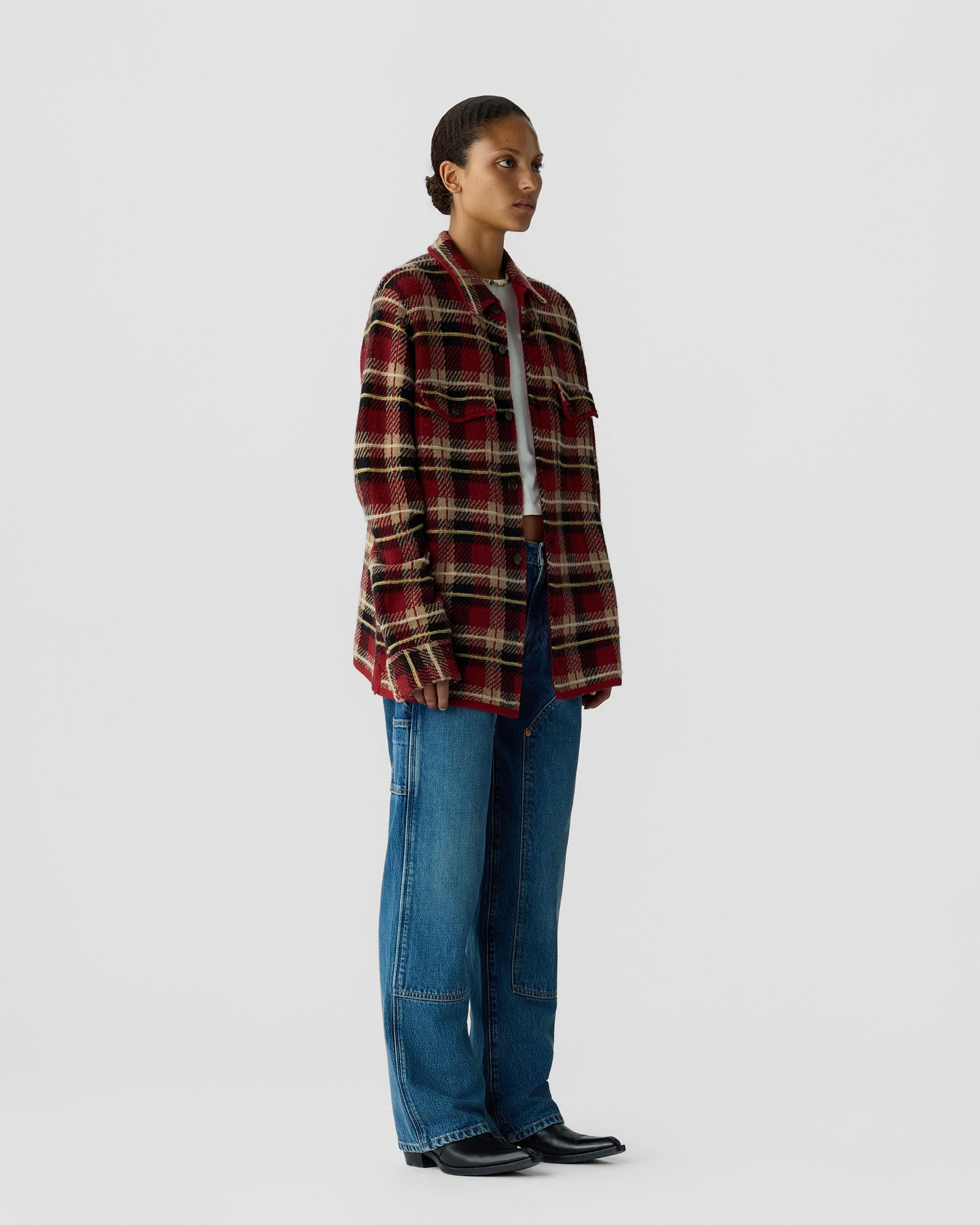 Profile view of a person wearing a red and beige plaid overshirt, light gray top, loose blue jeans, and black pointed shoes against a plain light gray background.