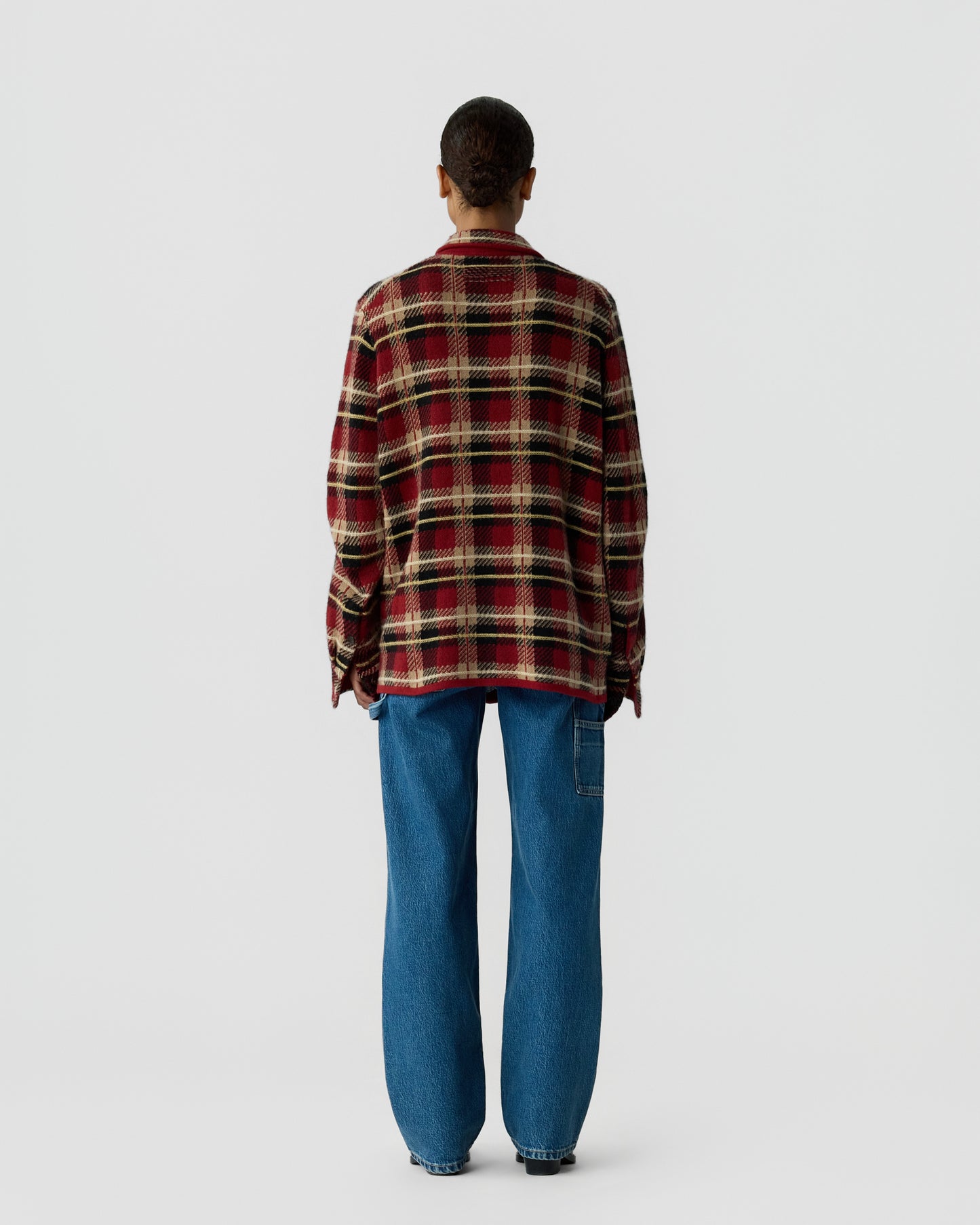 Person standing facing away wearing a red and beige plaid long-sleeve shirt and loose blue jeans against a plain light gray background.