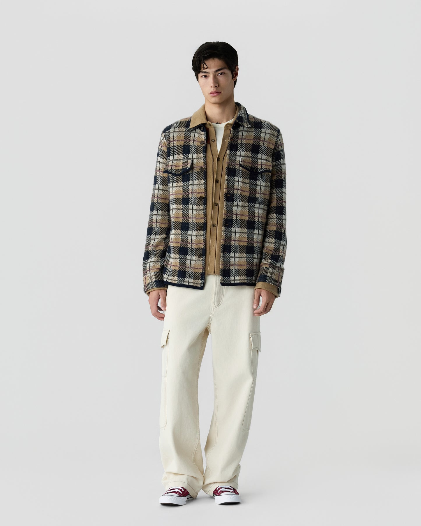 A young man standing against a plain gray background, wearing a brown and navy plaid jacket over a light brown shirt, cream cargo pants, and white sneakers with red details.