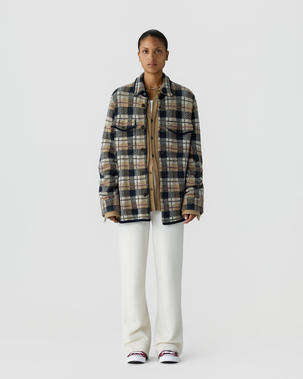 A person standing against a plain light gray background wearing a beige and navy plaid jacket over a tan buttoned shirt, white wide-leg pants, and white sneakers with red accents.