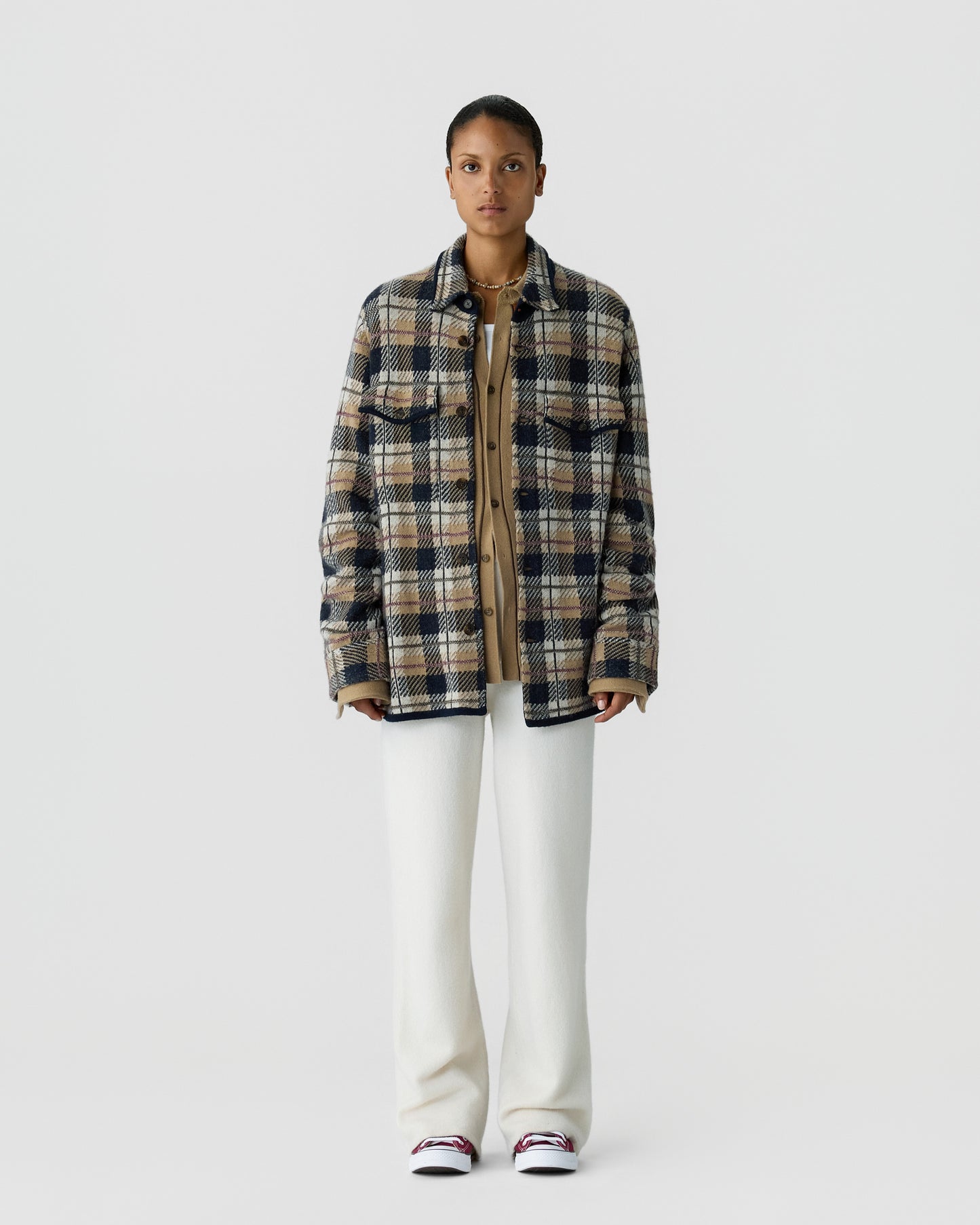 A person standing against a plain light gray background wearing a beige and navy plaid jacket over a tan buttoned shirt, white wide-leg pants, and white sneakers with red accents.