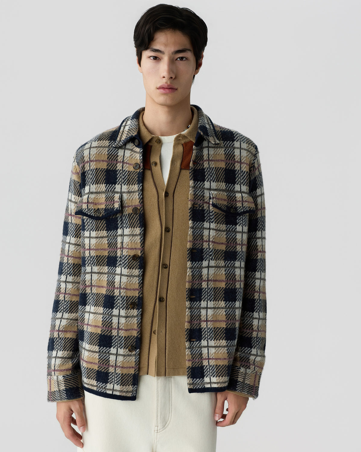 Young man wearing a beige and navy plaid jacket layered over a camel-colored button-up cardigan and a white undershirt, standing against a plain light background.