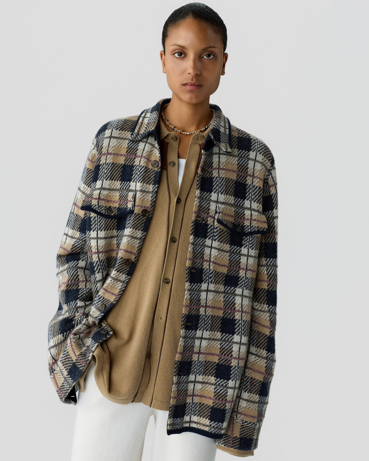 Woman wearing a beige button-up shirt layered under an oversized plaid jacket in navy, beige, and white tones, paired with white pants against a plain background.