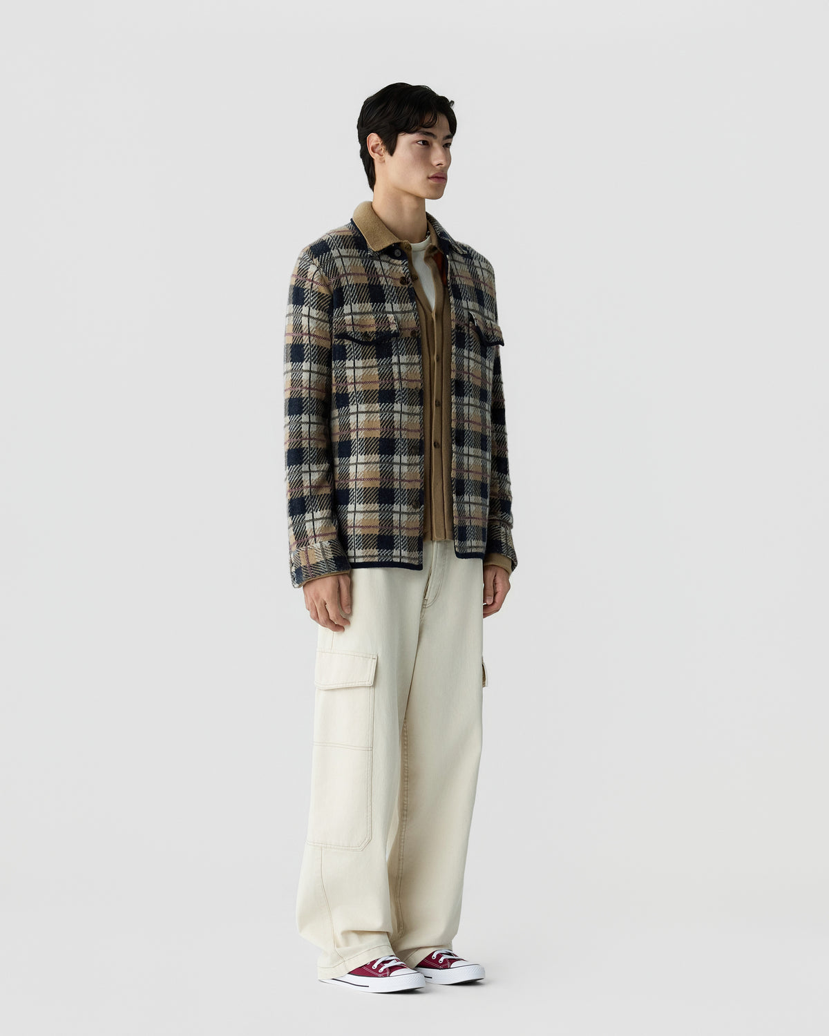 A man stands against a plain background wearing a plaid checkered jacket over a brown shirt, paired with wide cream cargo pants and maroon and white sneakers.