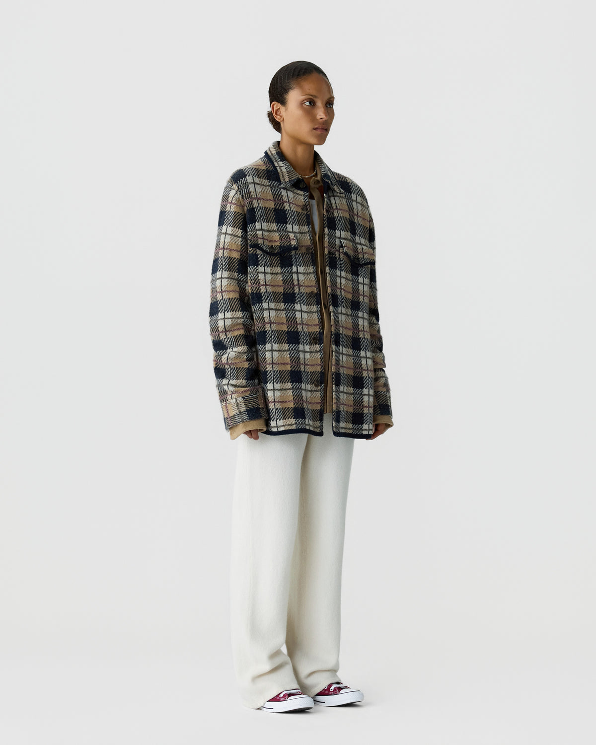 Woman wearing a plaid jacket with blue, beige, and brown tones over a brown top paired with loose white pants and red and white sneakers, standing against a plain white background.