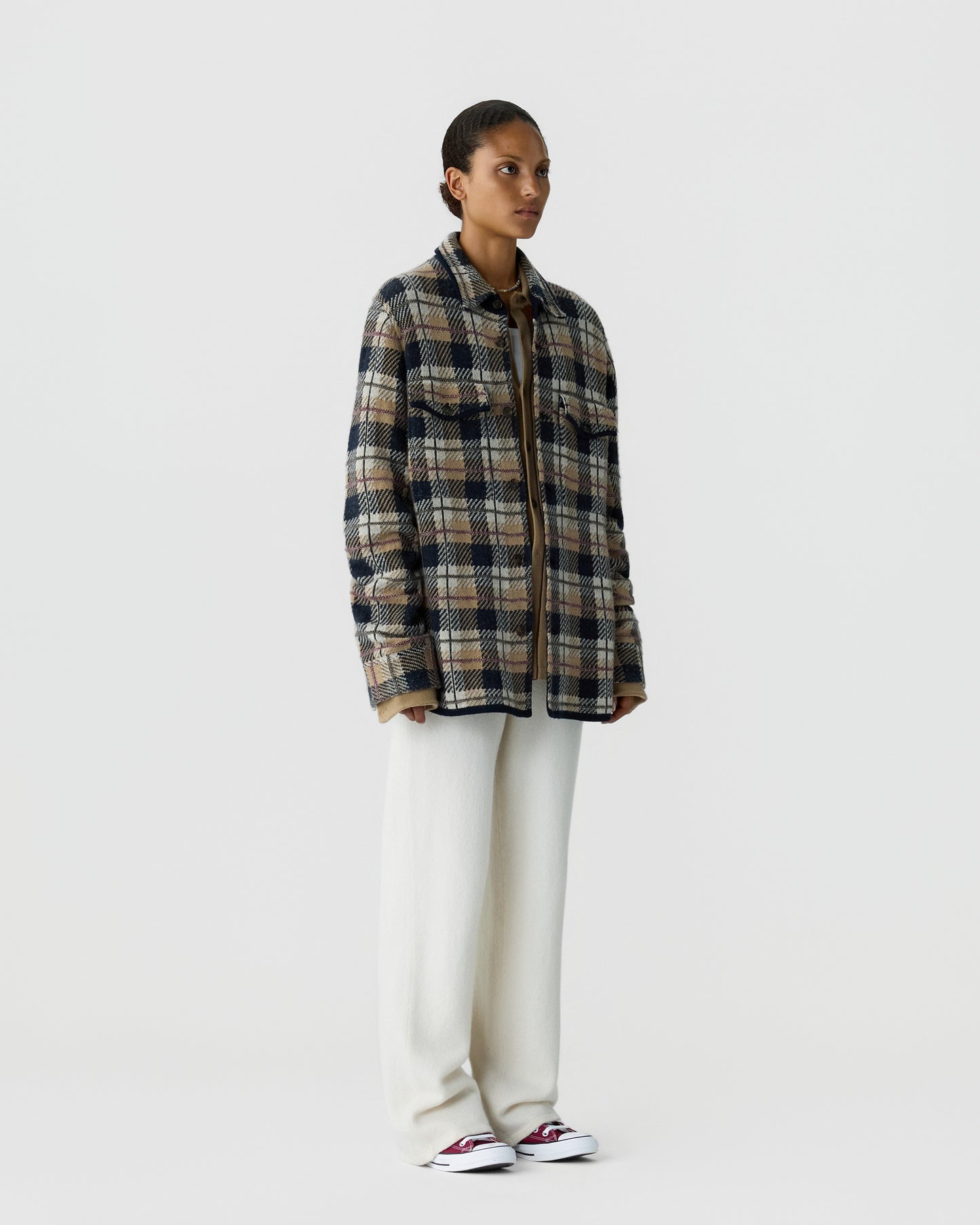 Woman wearing a plaid jacket with blue, beige, and brown tones over a brown top paired with loose white pants and red and white sneakers, standing against a plain white background.