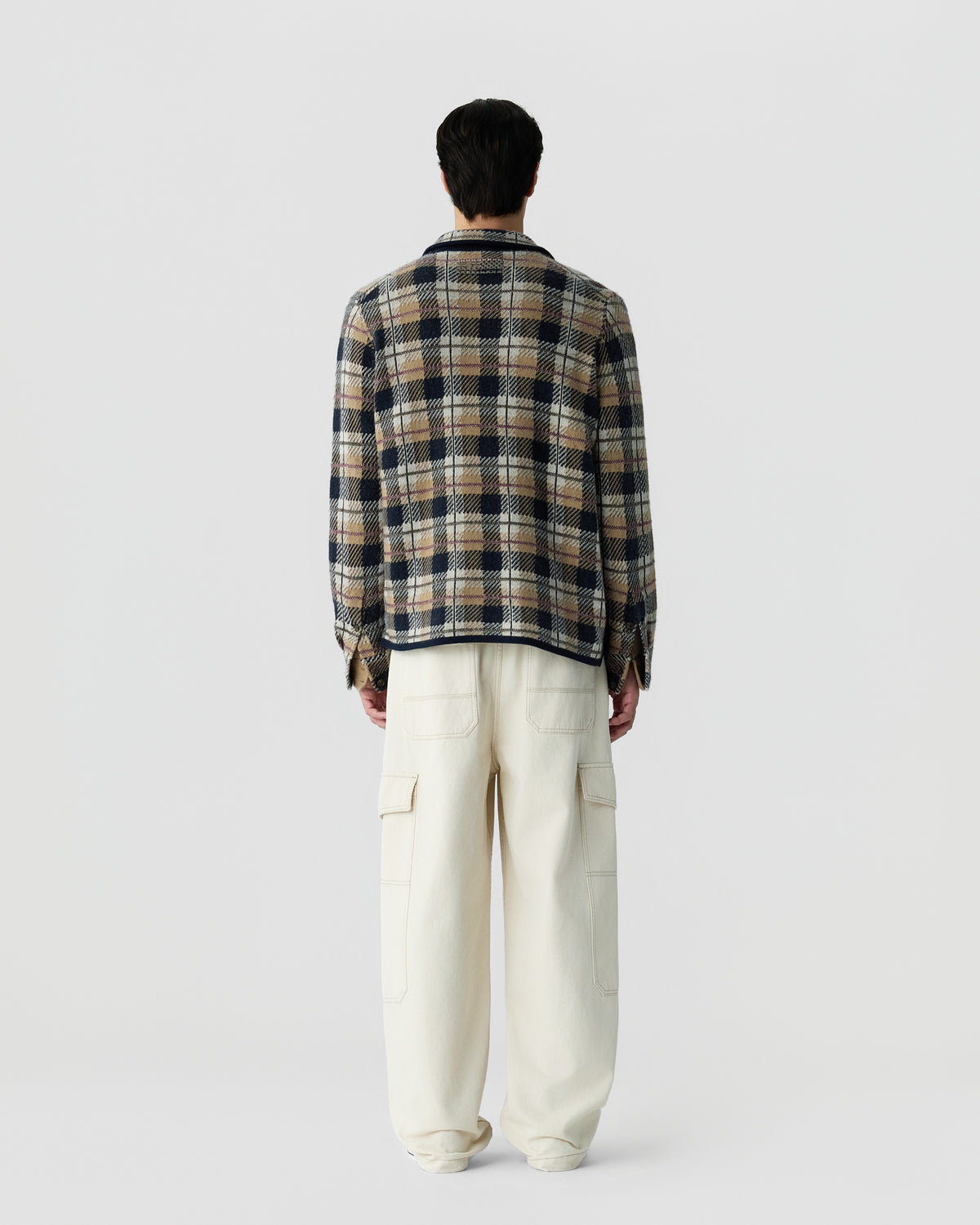 Man wearing a long-sleeve plaid shirt in brown, navy, and cream tones, paired with loose-fitting off-white cargo pants, standing facing away against a plain background.