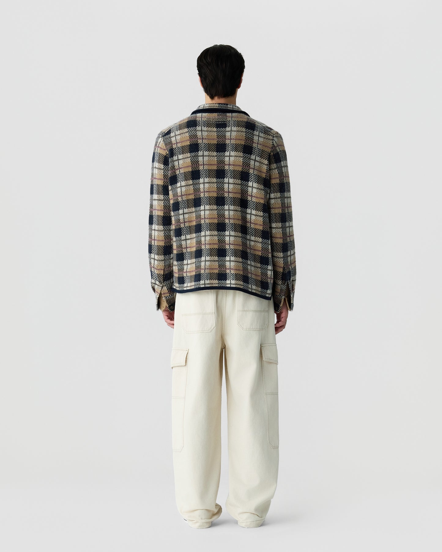 Man wearing a long-sleeve plaid shirt in brown, navy, and cream tones, paired with loose-fitting off-white cargo pants, standing facing away against a plain background.