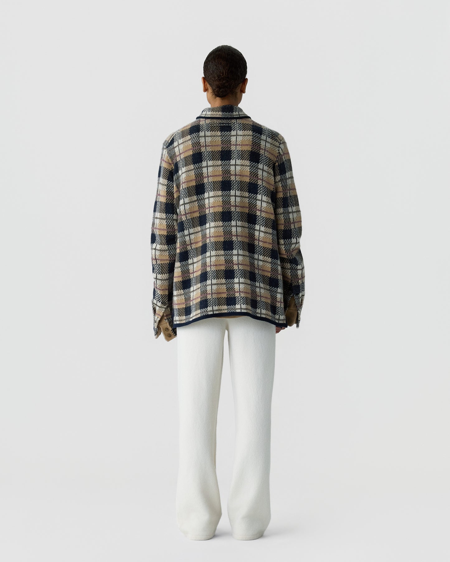 Person standing facing away wearing a loose plaid jacket in shades of brown, navy, and cream, paired with wide white pants against a plain light background.