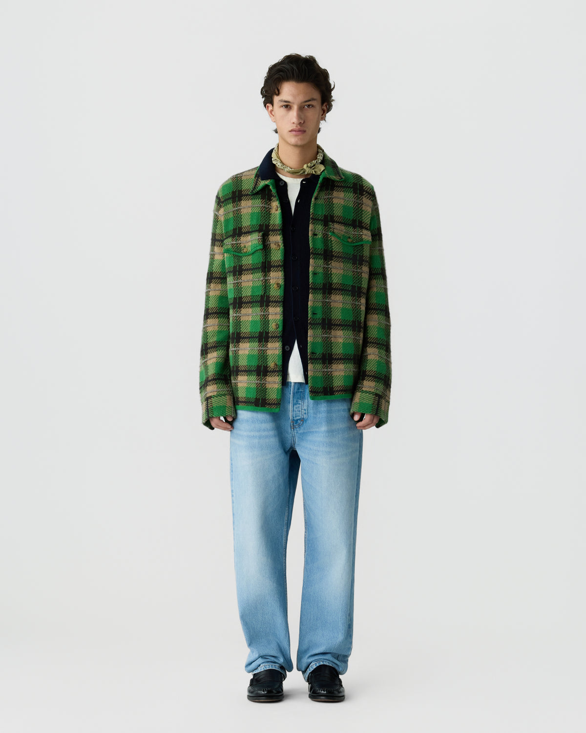 A young man wearing a green and black plaid jacket over a white shirt and layered with a black zip-up underneath, paired with light blue jeans and black shoes, standing against a plain white background.