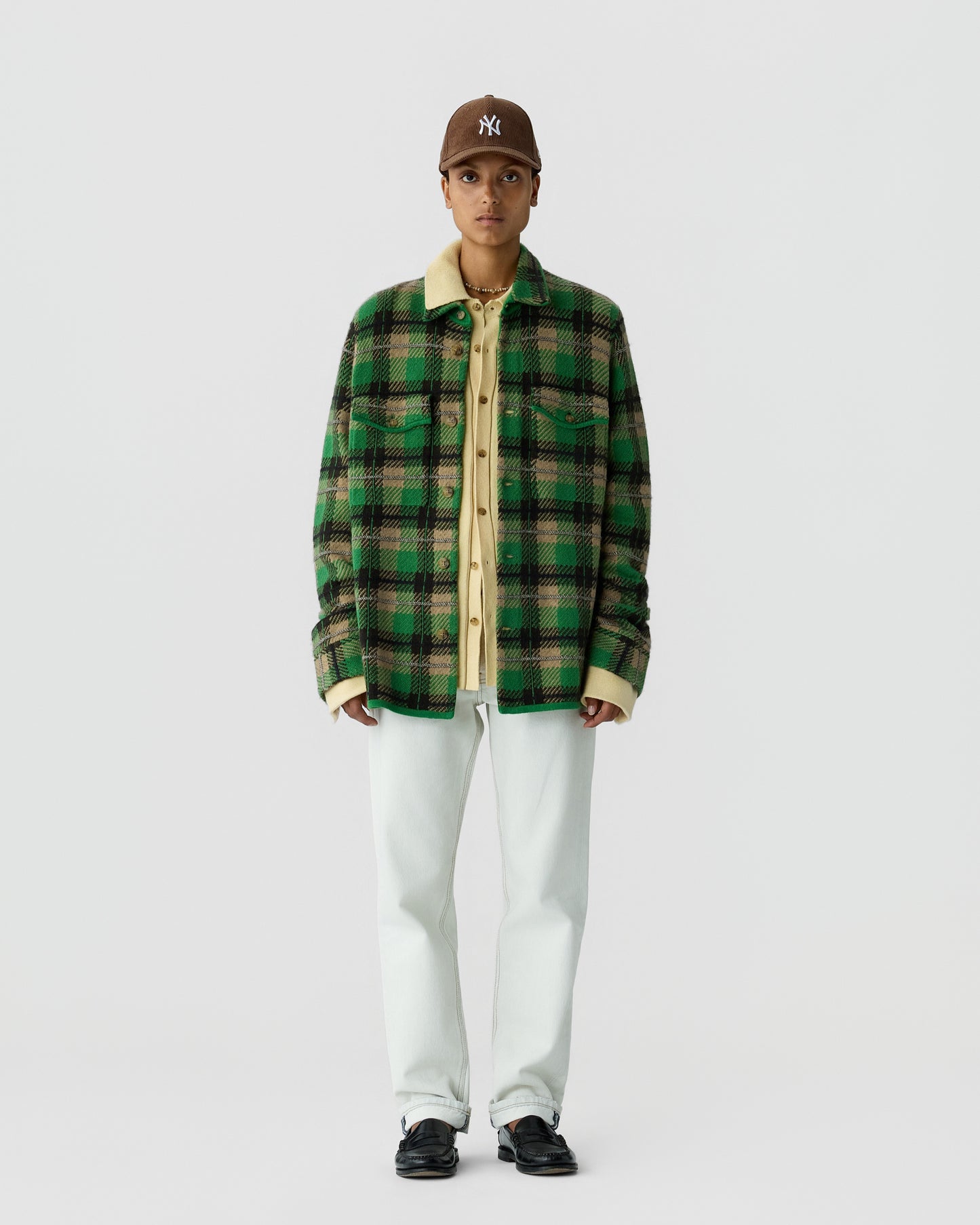 Person wearing a brown baseball cap, green and black plaid jacket with a beige collar and cuffs, beige shirt underneath, white pants, and black shoes, standing against a plain background.
