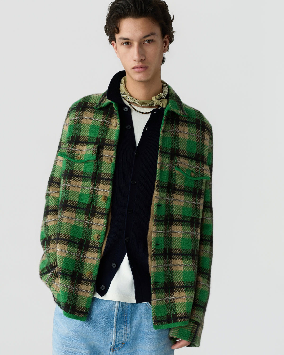 Young man wearing a green and beige plaid jacket over a black buttoned cardigan and white shirt, paired with light blue jeans and a patterned neck scarf.