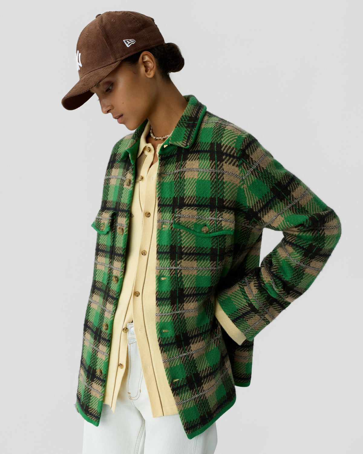 Woman wearing a green and black plaid jacket over a pale yellow button-up shirt paired with white pants and a brown baseball cap, standing against a plain background.