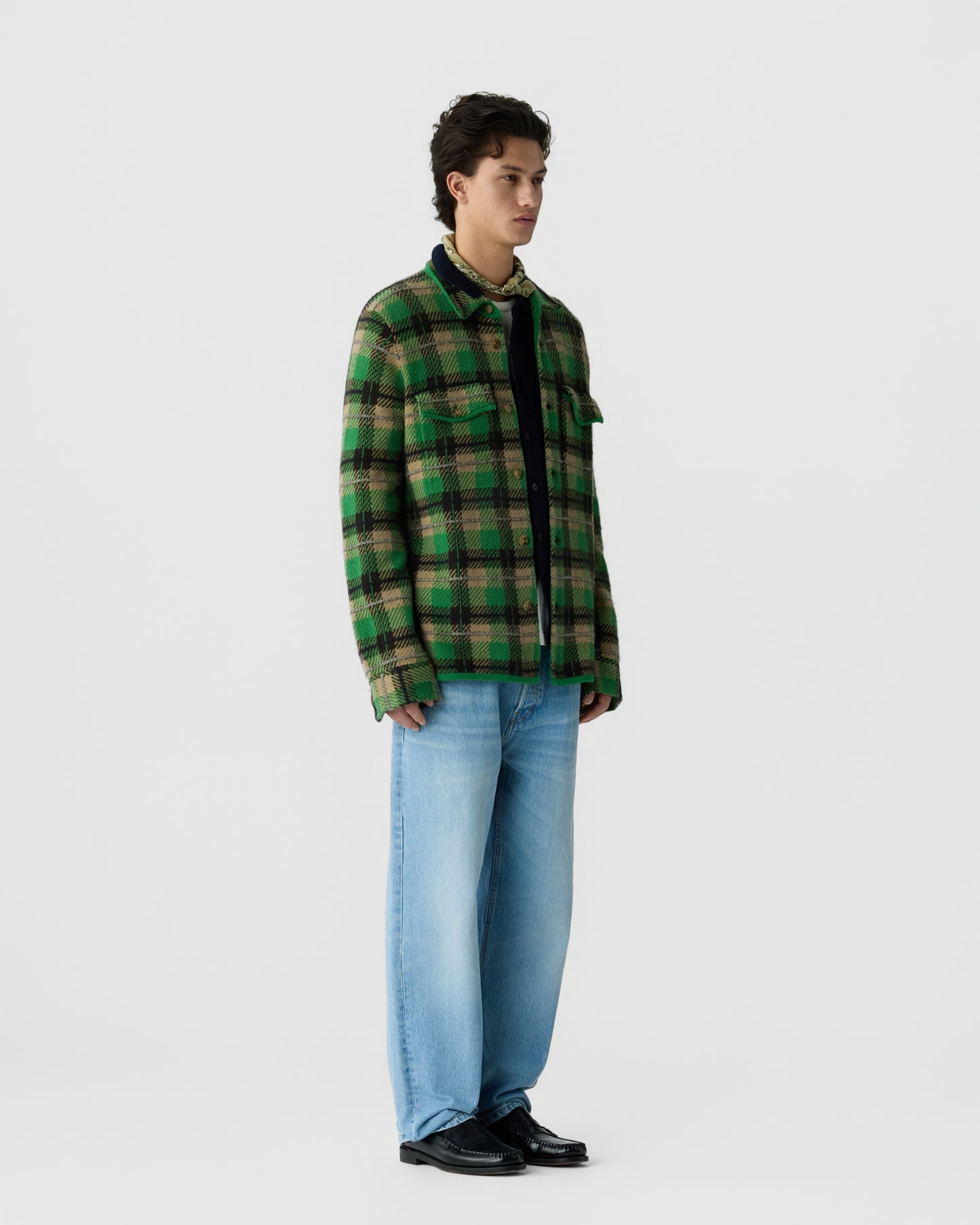 Young man wearing a green and black plaid button-up shirt with light blue wide-leg jeans and black shoes standing against a plain white background.