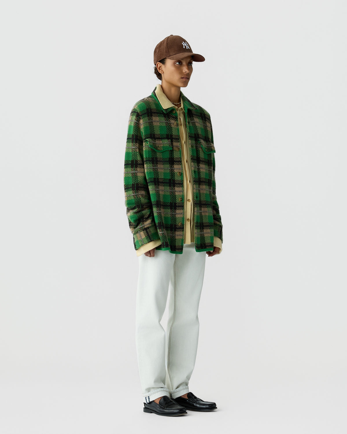 Person wearing a green and black plaid jacket over a beige shirt, white pants, black shoes, and a brown baseball cap standing against a plain light gray background.
