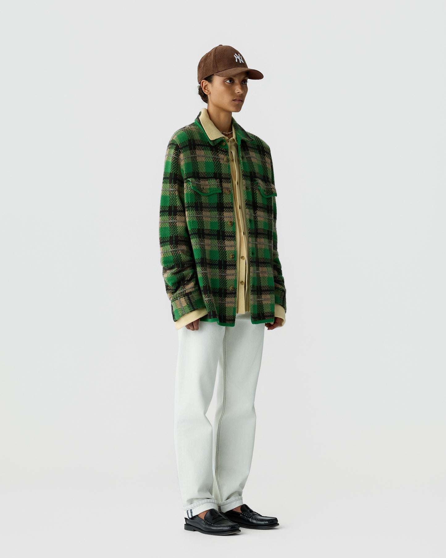 Person wearing a green and black plaid jacket over a beige shirt, white pants, black shoes, and a brown baseball cap standing against a plain light gray background.