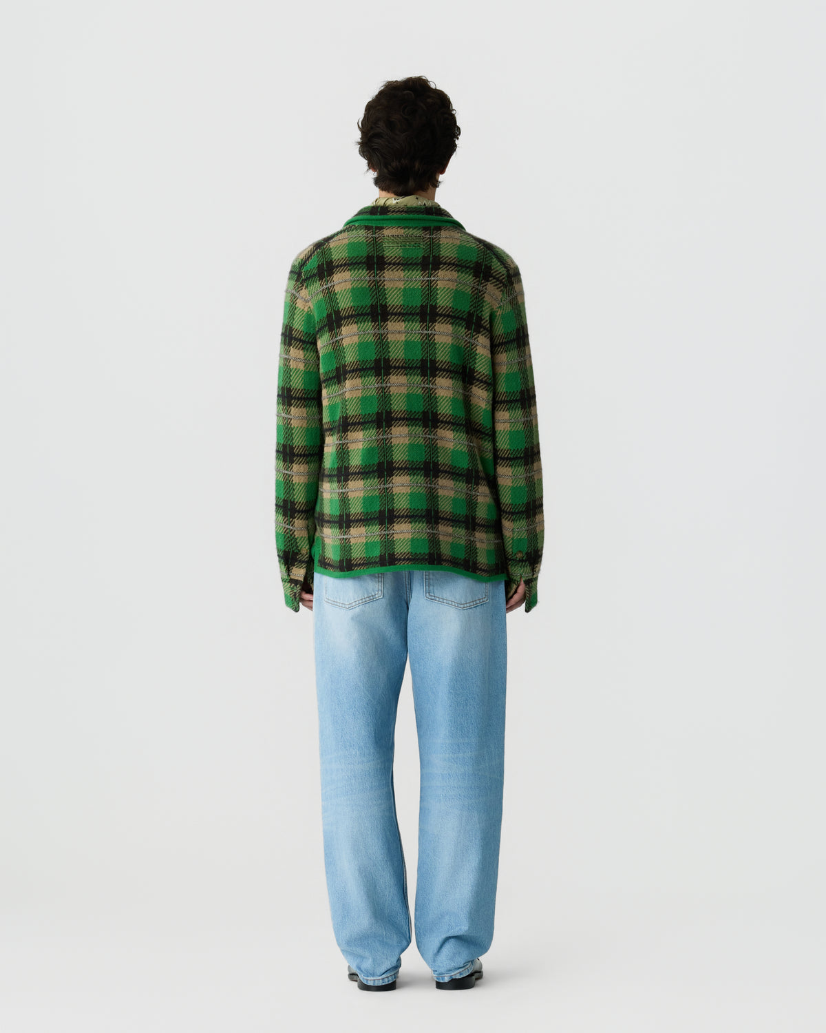 Person wearing a green and black checkered jacket and light blue jeans standing facing away against a plain white background.